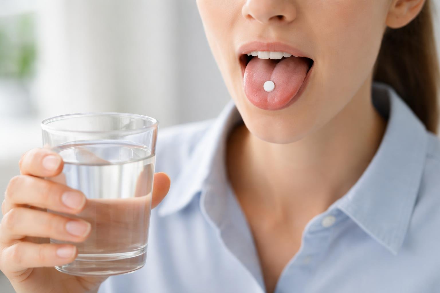 Pessoa segurando um comprimido sublingual na língua com um copo de água próximo sobre uma superfície branca.
