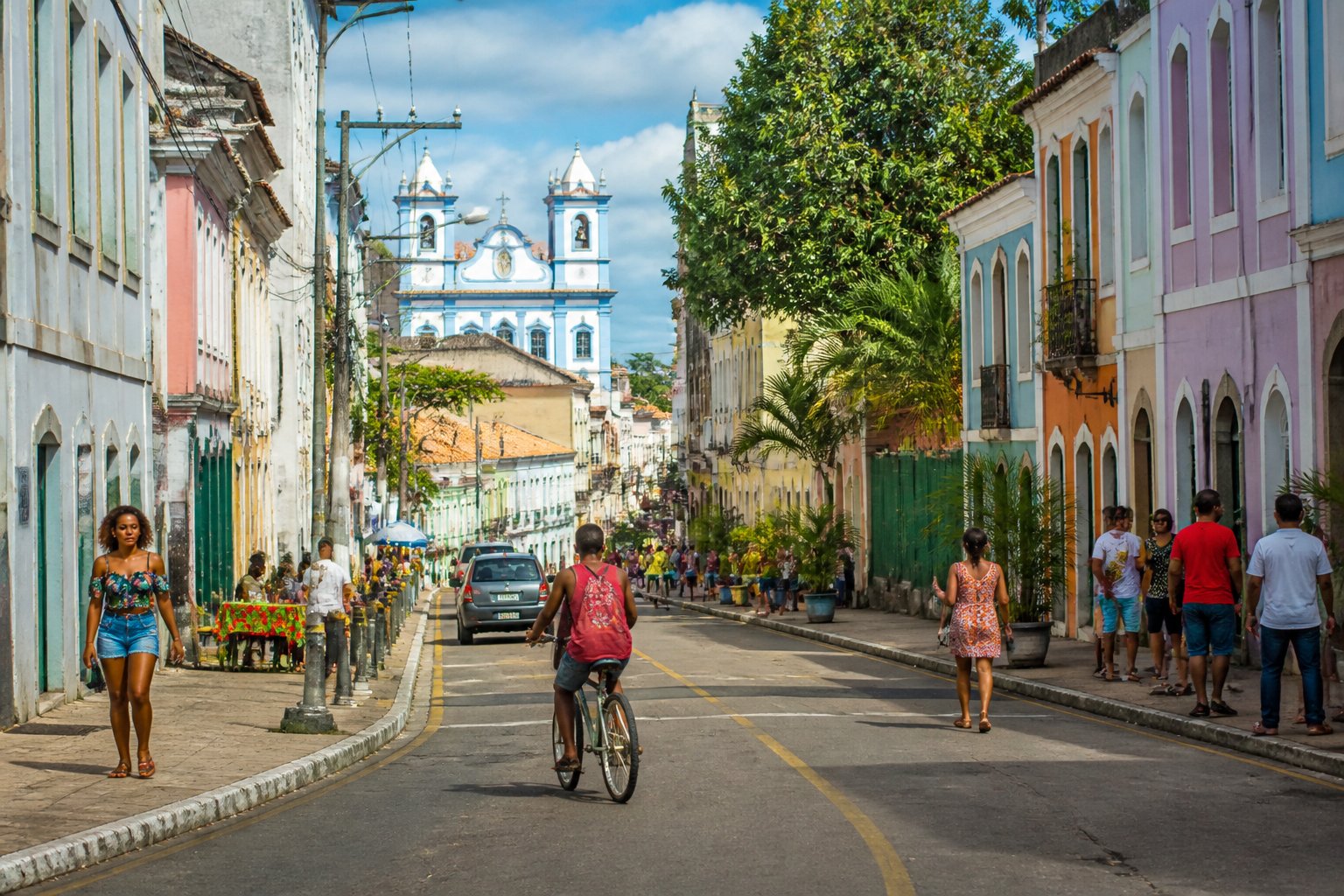 Cena de rua urbana no Brasil com pessoas caminhando e prédios coloridos ao fundo.