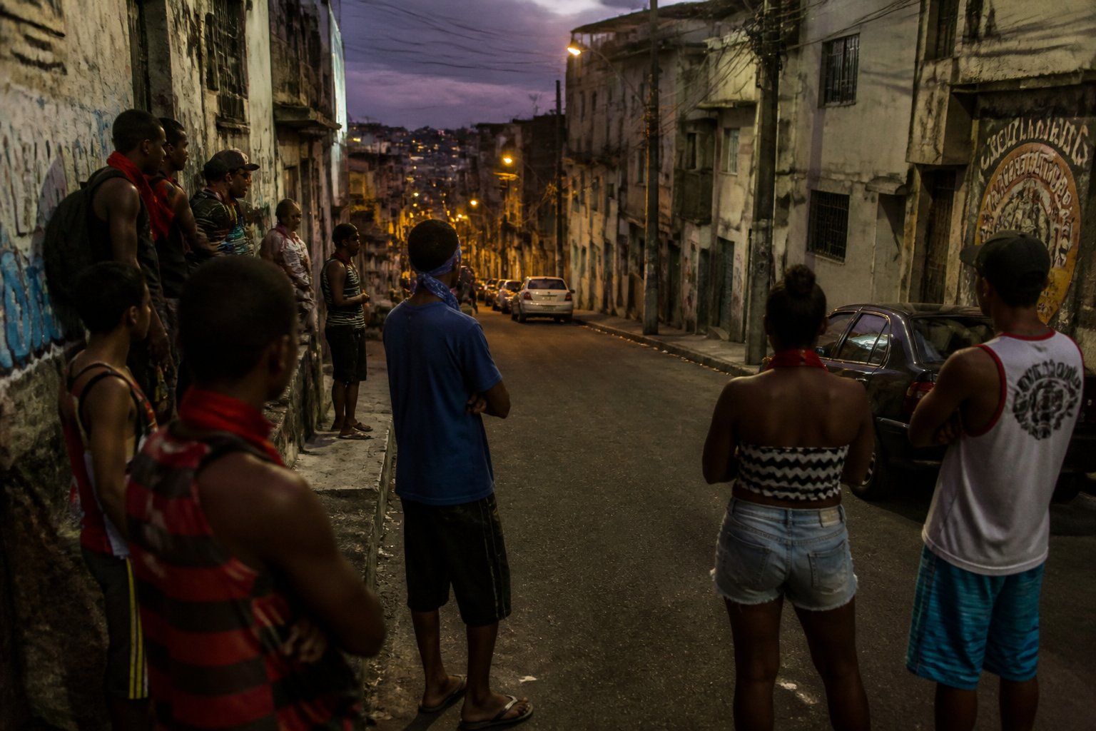 Rua estreita e mal iluminada em um bairro de Salvador com um grupo de jovens atentos e vigilantes, cercados por prédios desgastados e grafites.