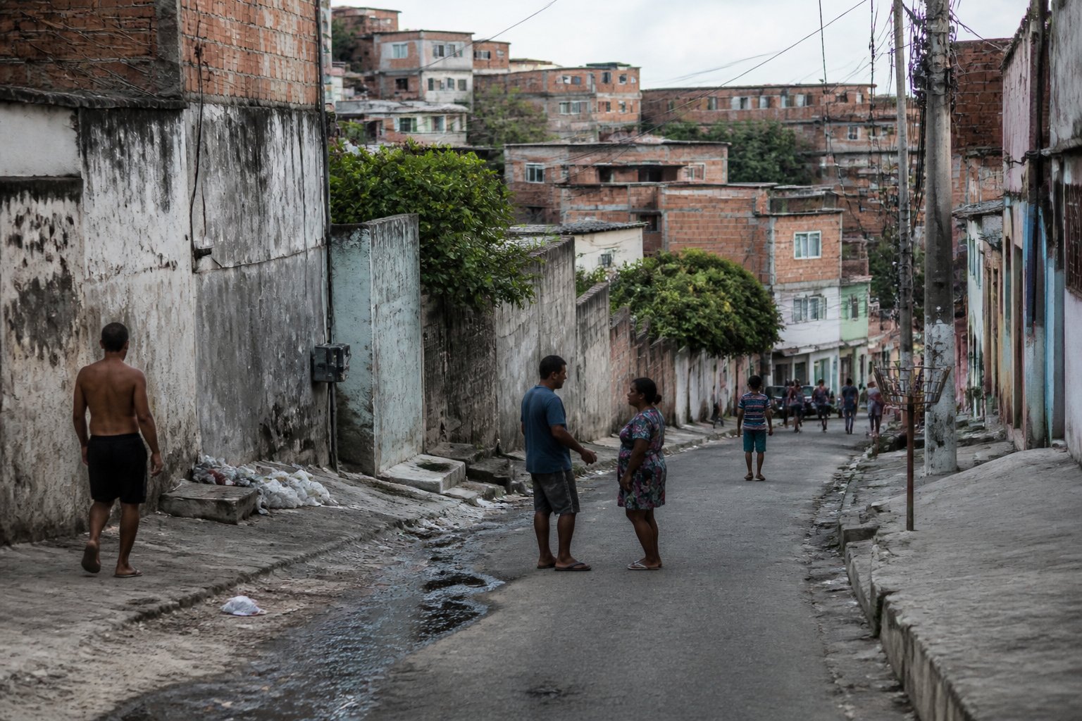 Vista de um bairro urbano em Cabo Frio com casas simples, ruas estreitas e algumas pessoas caminhando, mostrando sinais de dificuldades sociais.