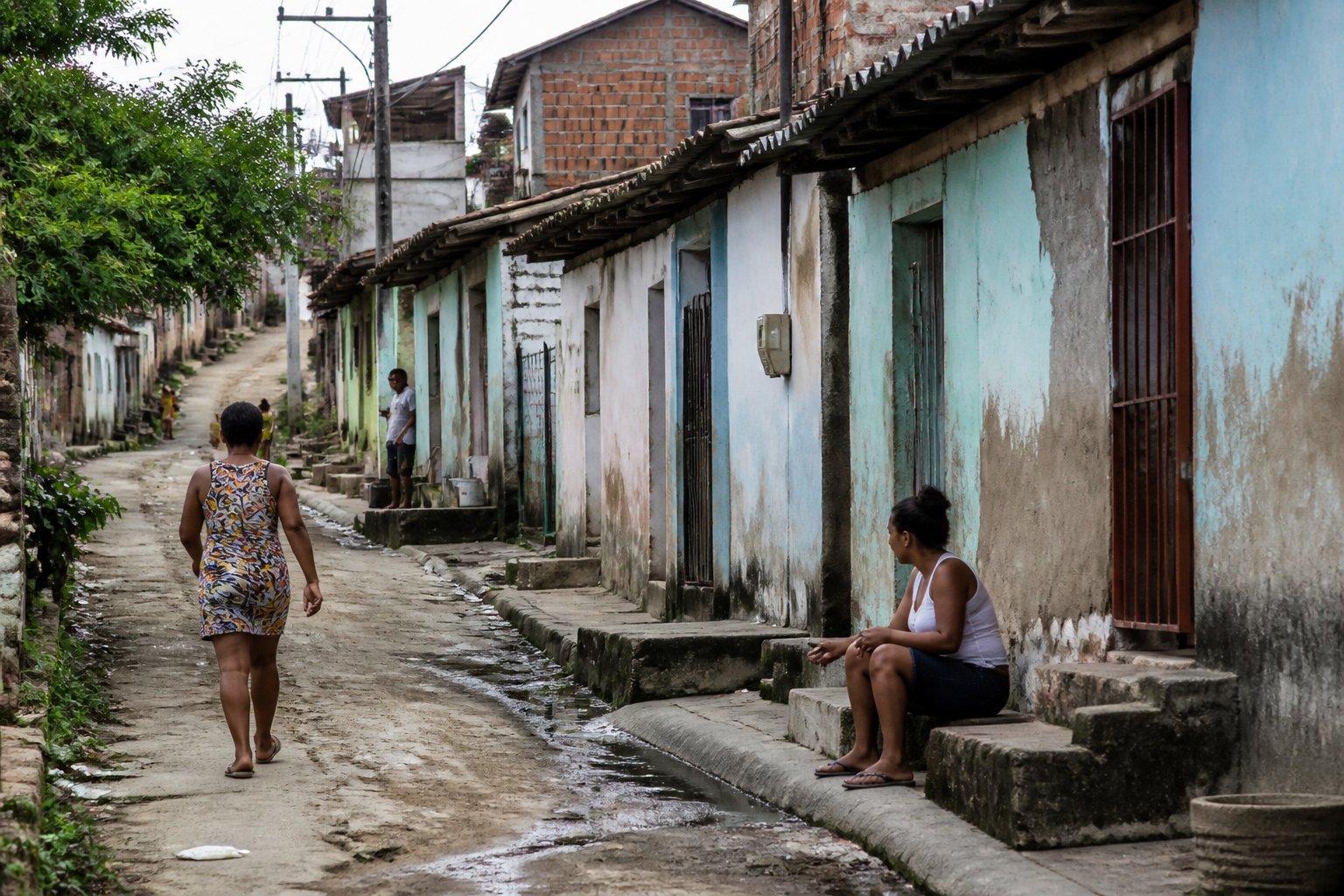 Rua de um bairro simples em João Pessoa com casas modestas, ruas estreitas e algumas pessoas caminhando.