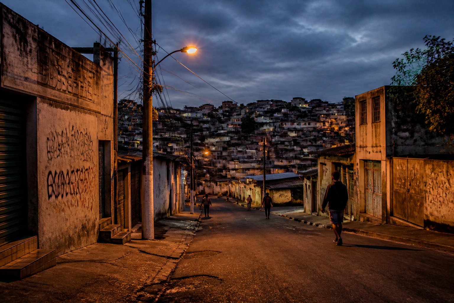 Rua de bairro urbano em Campinas ao anoitecer com iluminação fraca e algumas pessoas caminhando cautelosamente.