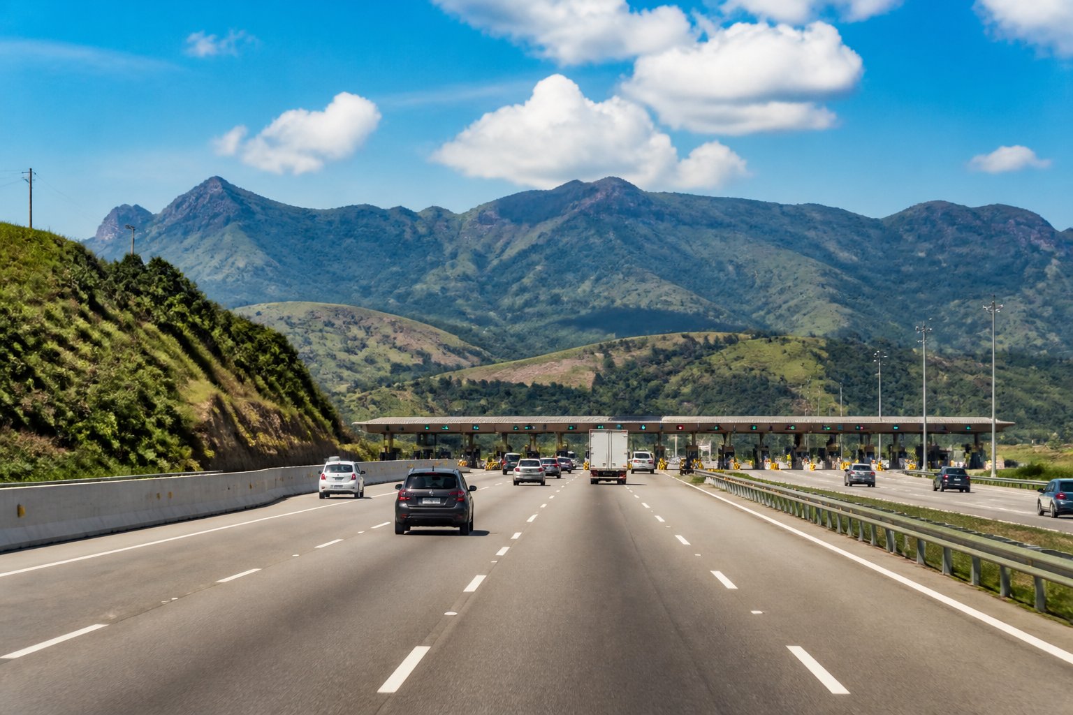 Rodovia brasileira com pedágios e veículos passando, cercada por vegetação e montanhas ao fundo.